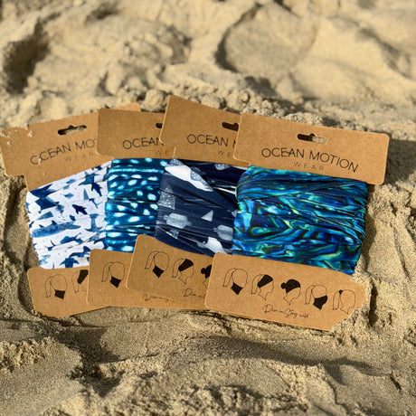Four packaged fabric headbands from Ocean Motion Wear are arranged on sand. The headbands feature blue, navy, and white patterns, including abstract swirls, spots, and ocean animal prints.