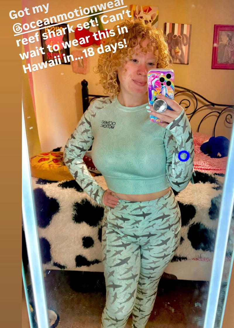 Womanwearing a green crop top and leggings with sharks and with text about ocean motionwear and Hawaii.