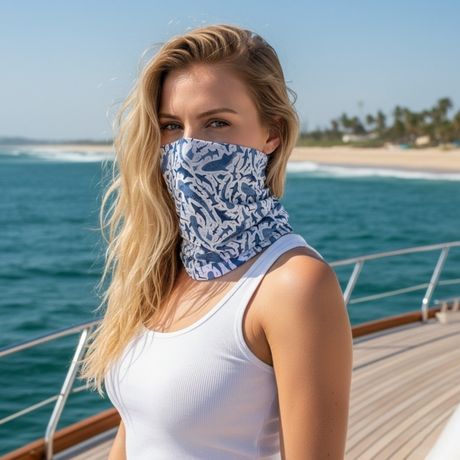 A woman on a yacht wearing a blue and white shark patterned neck gaiter, with the ocean and beach in the background.