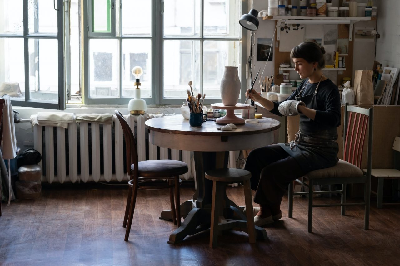 Young Woman In Apron Sits On Chair At Round Table Painting Handmade Ceramic Vase In Workshop.jpg